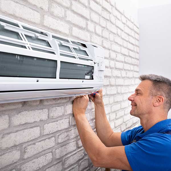 Man in a blue polo shirt installs or repairs a wall-mounted air conditioner on a brick exterior wall.