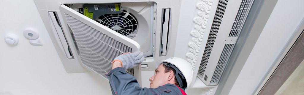Maintenance worker in a hard hat services a ceiling-mounted air conditioner unit while wearing gloves and a gray coverall; the unit's access panel is open.