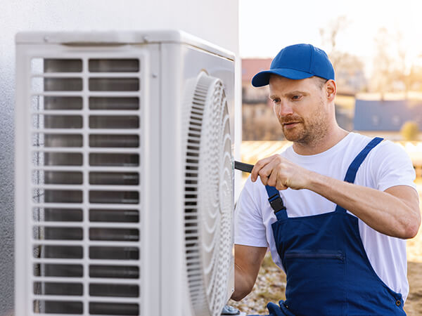 HVAC technician in a blue cap and overalls tightening a screw on an outdoor air conditioner unit