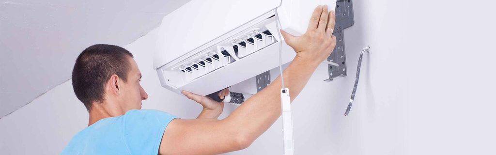 Technician mounts a wall-mounted air conditioner, securing the unit to the wall with hands visible and wiring nearby.