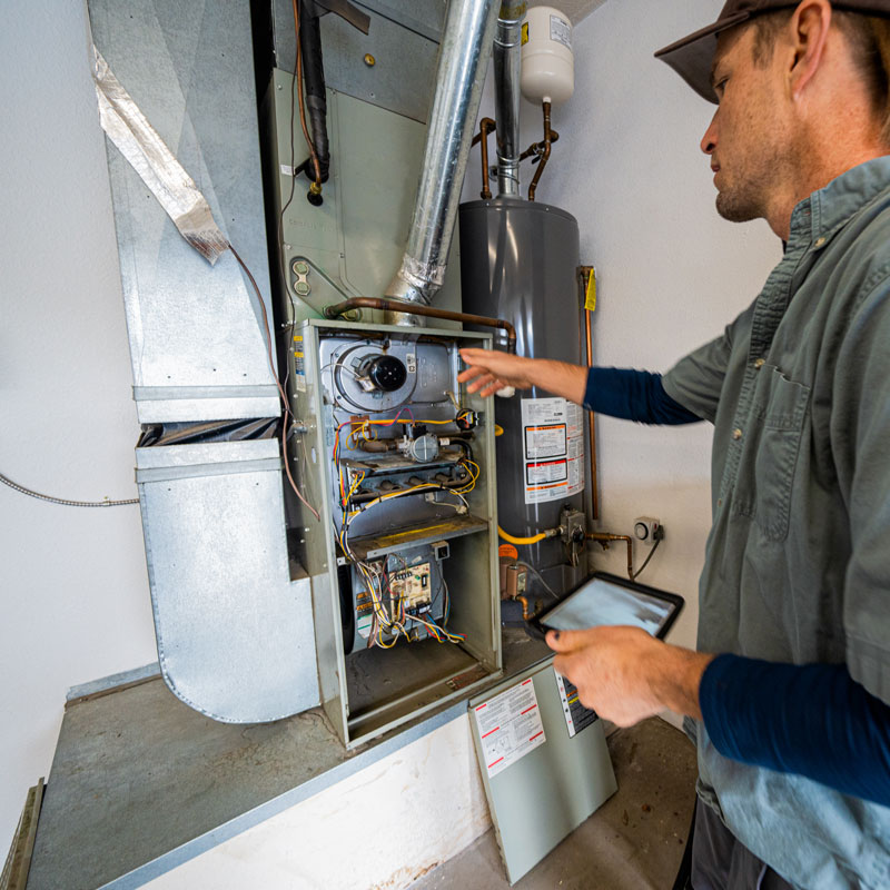 HVAC technician inspecting an open furnace control panel with exposed wiring, holding a tablet for diagnostics