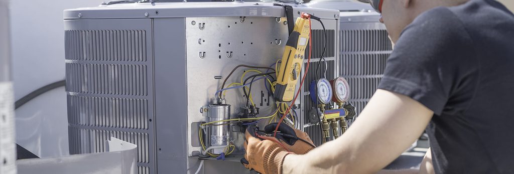 HVAC technician in gloves working on an outdoor air conditioner condenser, using a multimeter and manifold gauges to test connections.