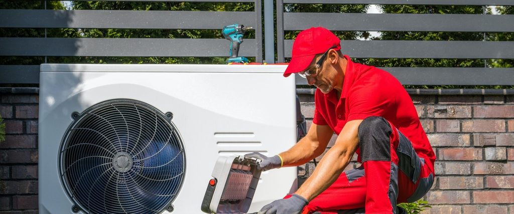 HVAC technician in a red uniform crouches by an outdoor air conditioner, inspecting the unit with a handheld tester; drill on the unit nearby.