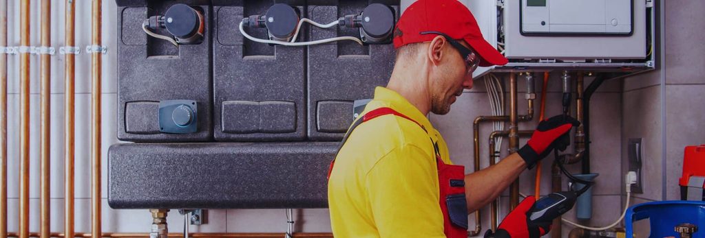 HVAC technician in a yellow uniform and red cap repairing a large boiler or furnace with copper pipes nearby.