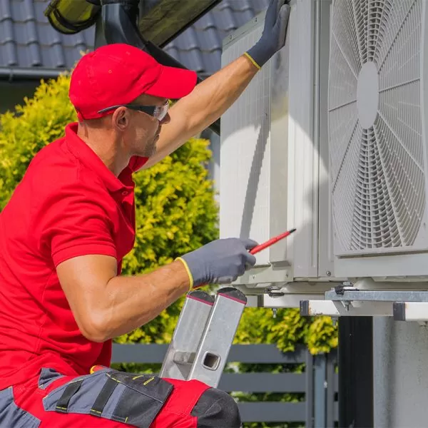 HVAC technician in a red uniform and cap using a screwdriver on an outdoor air conditioning condenser unit.